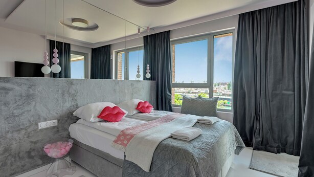 A modern bedroom with a large bed, a bedside table with a pink bowl, and a city view through the window. - Żurawie Pink Port by Grand Apartments (Gdańsk)