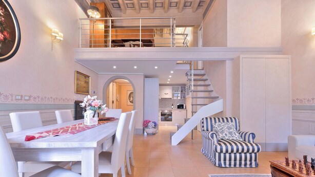 A spacious living area with a dining table, chairs, a striped armchair, and a chess table. A staircase leads to an upper level with a balcony. - Venus apartment (Firenze)