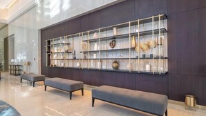 A modern lobby with a dark wood panel wall, a glass shelving unit displaying decorative items, and two grey upholstered benches.