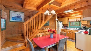 Interior de una cabaña de madera con una mesa de comedor, sillas, una zona de cocina y una escalera.