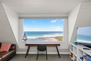 A room with a desk and chair facing a large window overlooking a beach and ocean.