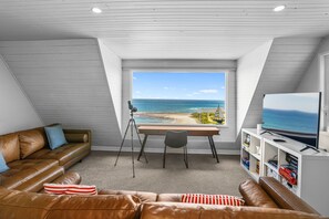 Ein gemütliches Wohnzimmer mit einem großen Fenster mit Blick auf einen Strand, ein Ledersofa, ein Schreibtisch mit Stuhl und ein Fernseher auf einem weißen Schrank.