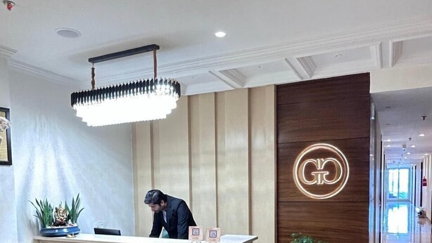 A reception desk with a modern chandelier, a wooden accent wall with a circular logo, and a potted plant. - GREEN GARDEN CAFE & HOTEL (Amritsar)