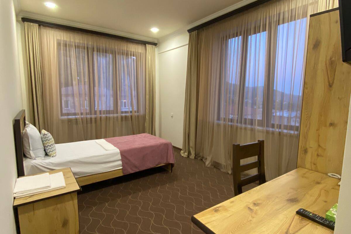Habitación de hotel con cama, escritorio y una silla. Hay cortinas en las ventanas y un televisor montado en la pared.