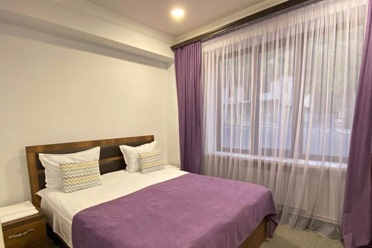 Un dormitorio con cama, mesita de noche, ventana con cortinas translúcidas y vista al exterior.