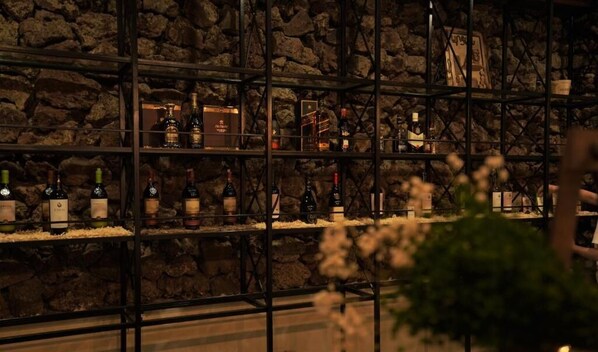 A bar area with shelves of bottles, a stone wall background, and a plant in the foreground. - Mina Hotel (Goris)