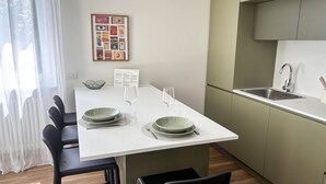 A modern dining area with a white table set for two, a framed collage on the wall, and a kitchen with a green cabinet and stainless steel sink.