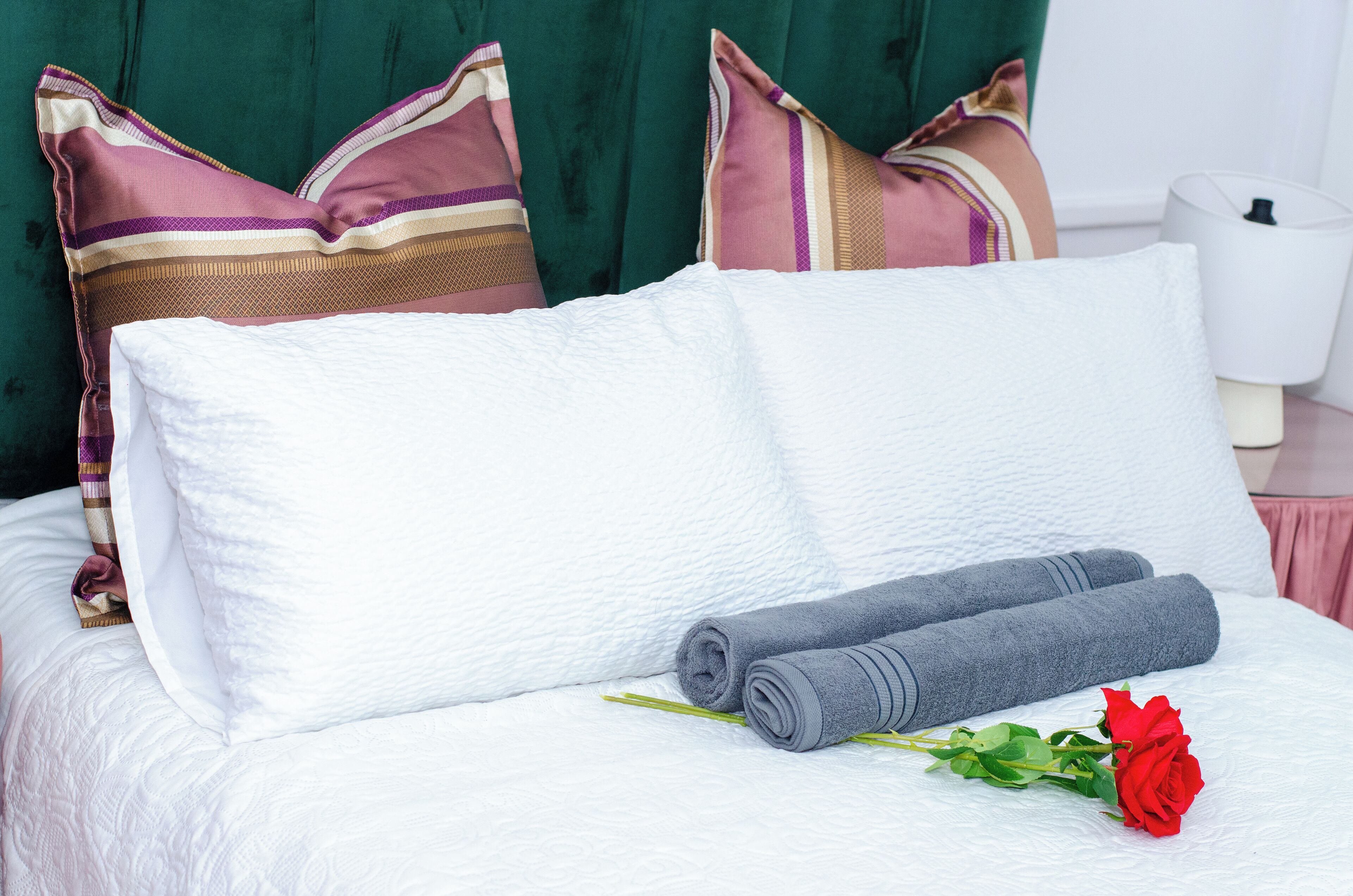 Una cama bien hecha con sábanas blancas, dos toallas grises enrolladas y una rosa roja.