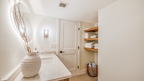 A bathroom with a white sink, a large vase with branches, a towel rack with folded towels, and a basket.