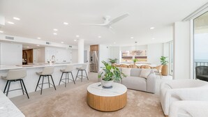 Una sala de estar moderna con una mesa de centro redonda de madera, un sofá y una zona de cocina con encimera blanca y taburetes de bar.
