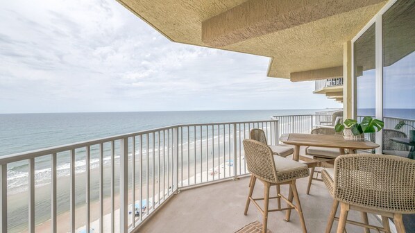 A balcony with a view of the beach and ocean. There are wicker chairs and a table with a plant.