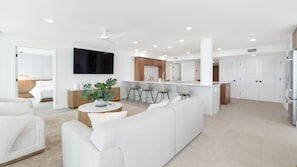 Una sala moderna con un sofá, una mesa de centro y un televisor. Se observa un área de cocina con una barra y taburetes.
