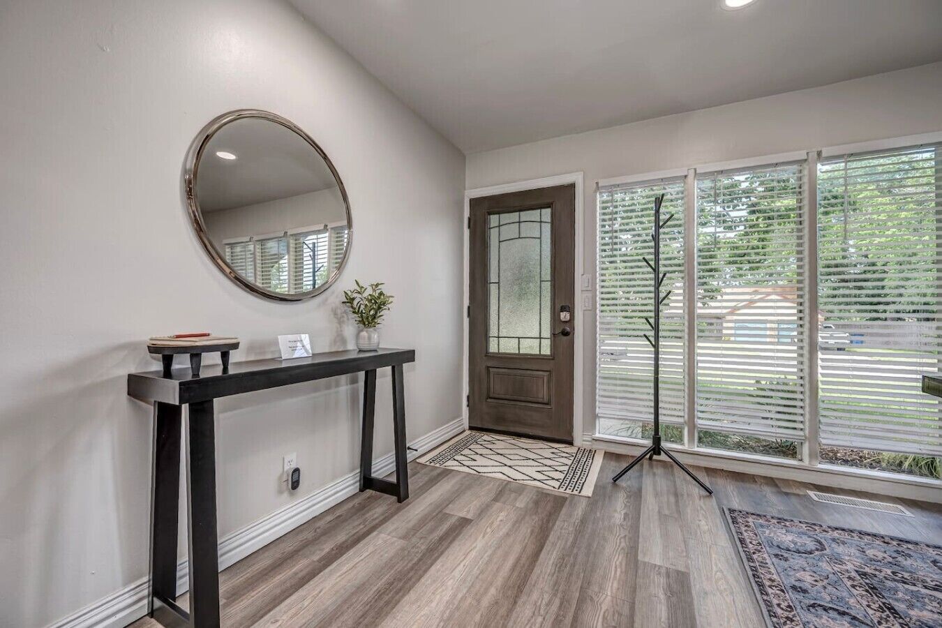 A modern entryway with a dark console table, a round mirror, a potted plant, a door with frosted glass panels, and a large window with blinds.