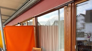 Una terraza de madera con una mesa, sillas, un ventanal corredero y vistas al exterior.
