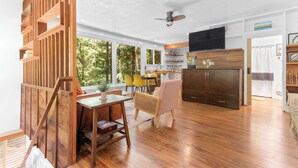 Un amplio salón con suelo de madera, una mesa de comedor con sillas, una zona de cocina con televisor y vistas a árboles a través de grandes ventanales.