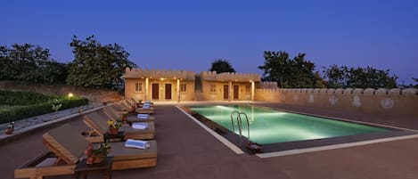 A poolside area with lounge chairs and a swimming pool, surrounded by a low wall and trees.