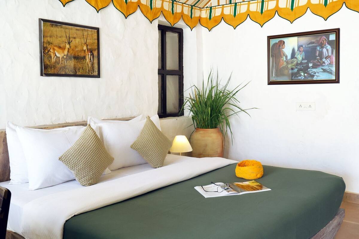 Un dormitorio con cama, almohadas, una lámpara, una planta y dos cuadros en la pared.