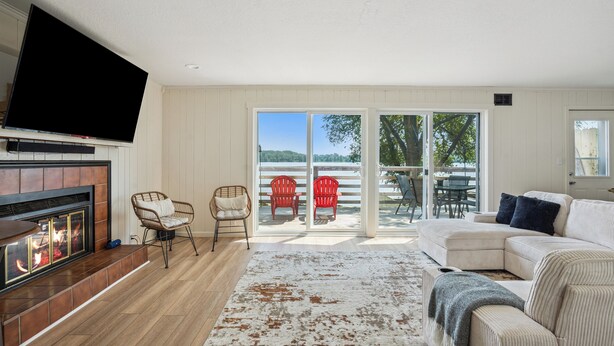 A living room with a fireplace, a flat-screen TV, a sectional sofa, and a large window with a view of trees and a lake. - The Big Muddy Riverfront Bungalow (Princeton)