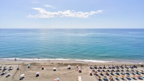 Une plage bordée de rangées de transats et de parasols, un rivage sablonneux et l’océan.