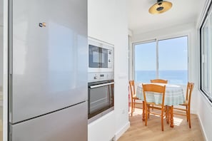 Una cocina compacta con refrigerador, horno y microondas, una mesa de comedor con sillas y un ventanal con vistas al mar.