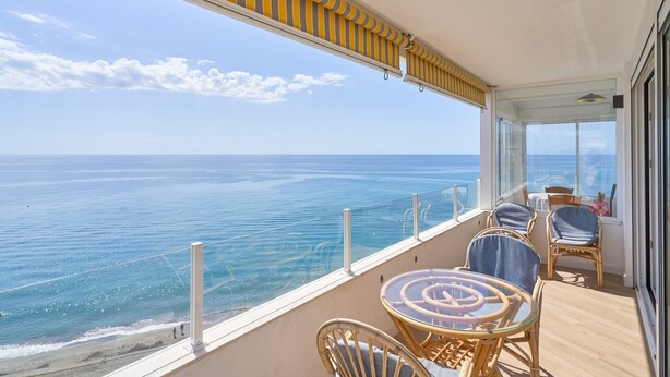 Un balcon avec vue sur l’océan, des meubles en osier et une porte vitrée.