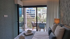 Habitació d'hotel amb llit, sofà, balcó amb taula i cadires, i vista a edificis i vegetació.