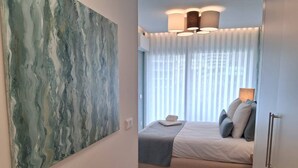 Un dormitori amb un llit, un quadre abstracte gran, una finestra amb cortines i un llum al sostre.