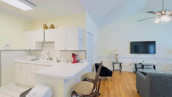 Une cuisine moderne avec des meubles blancs et un îlot central, un coin repas avec un canapé gris et une petite table, et un téléviseur fixé au mur.