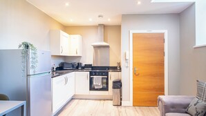 Una cocina moderna con armarios blancos, un refrigerador de acero inoxidable, un microondas y una estufa. Hay una puerta de madera con un letrero que dice 'No Smoking', un bote de basura y una pequeña mesa con una planta.