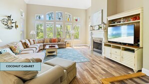 Una sala de estar con televisor grande, chimenea y vistas a árboles a través de las ventanas.