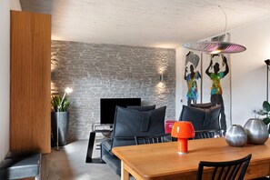 Ein modernes Wohnzimmer mit Steinwand, Flachbildfernseher, einer Couch, einem Esstisch und einer roten Lampe.