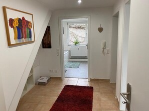 Un pasillo con un tapete rojo, un cuadro de flores y vista hacia un baño con una planta.