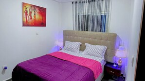 A bedroom with a bed, a nightstand, a lamp, a ceiling light, a window with curtains, and a painting on the wall.