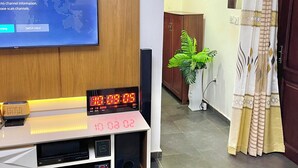 A modern living room with a flat-screen TV mounted on a wooden wall, a digital clock, and a small speaker system.