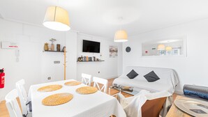 A modern, well-lit room with a dining table, chairs, a sofa, a TV, and a bed with pillows.