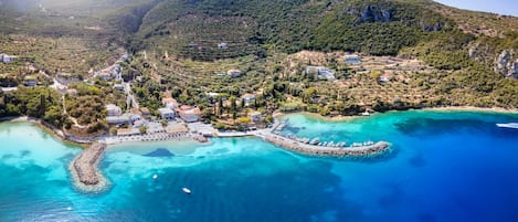 Vue aérienne d’une ville côtière, avec ses eaux turquoise et limpides, un brise-lames rocheux et des bâtiments entourés de verdure.
