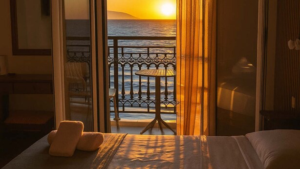 Une chambre d’hôtel avec un lit, une vue sur l’océan au coucher du soleil, un balcon avec une table et une chaise.