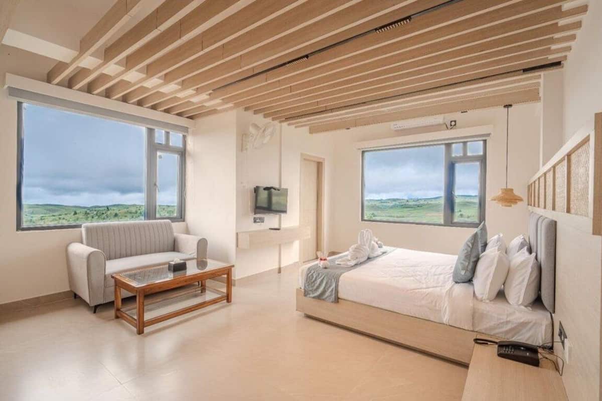 A modern bedroom with a large bed, a sofa, a TV, and large windows offering a view of the countryside.