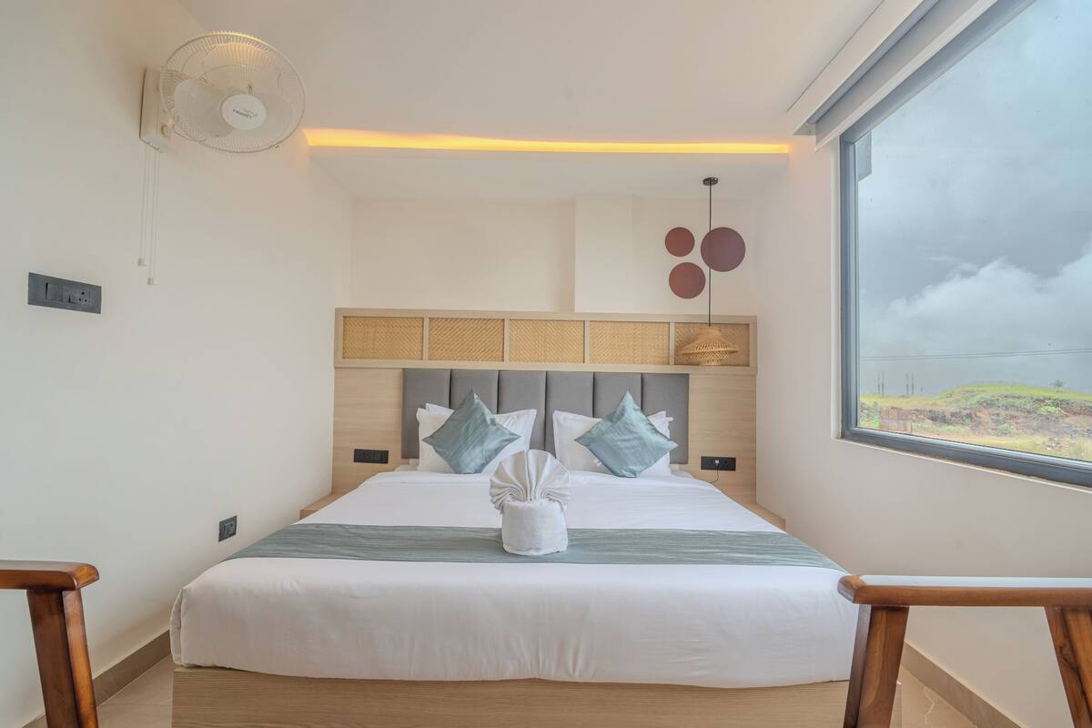 A modern hotel room with a large bed, a wooden headboard, a bedside table, a standing fan, and a large window with a view of the countryside.