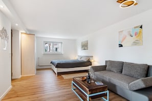 Une chambre moderne avec un lit, un canapé, une table basse en bois et une étagère murale ornée de livres et d’un vase.