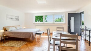 A bedroom with a bed, bedside tables, a window, a dining area with chairs, and a view of greenery outside.