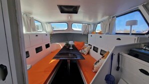 Interior de barco pequeño con una mesa central, cojines naranja y una ventana.