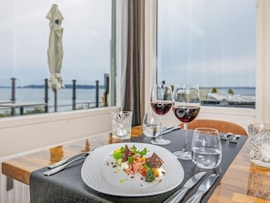 A dining table with a plate of food, two glasses of red wine, and a view of the sea through a window.