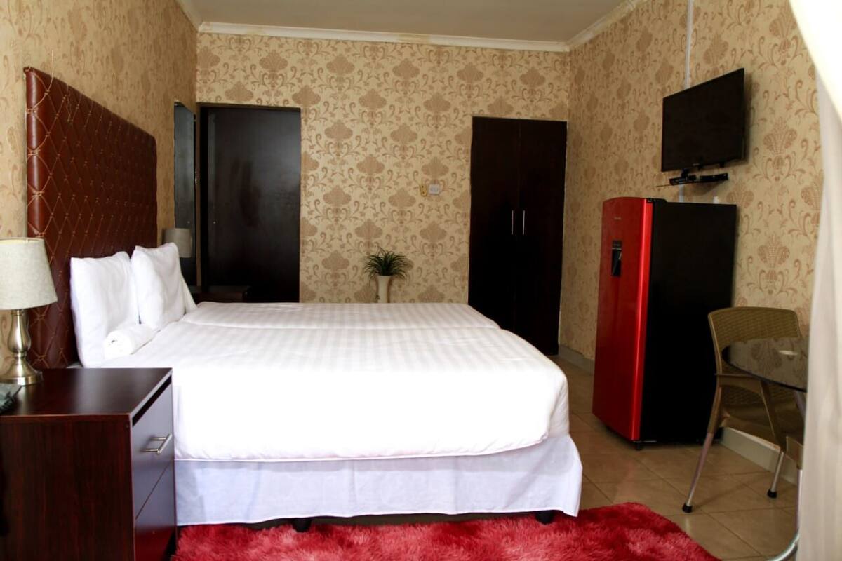 A hotel room with a bed, a desk with a lamp, a TV, and a red refrigerator.