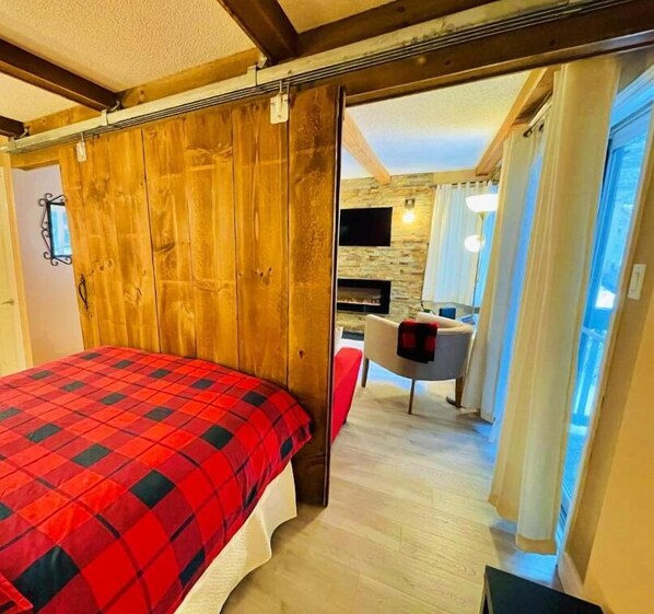 A bedroom with a wooden headboard, a red and black checkered bedspread, a television mounted on the wall, and a fireplace. - Rustic Mountainside Chalet (Blue Mountain)