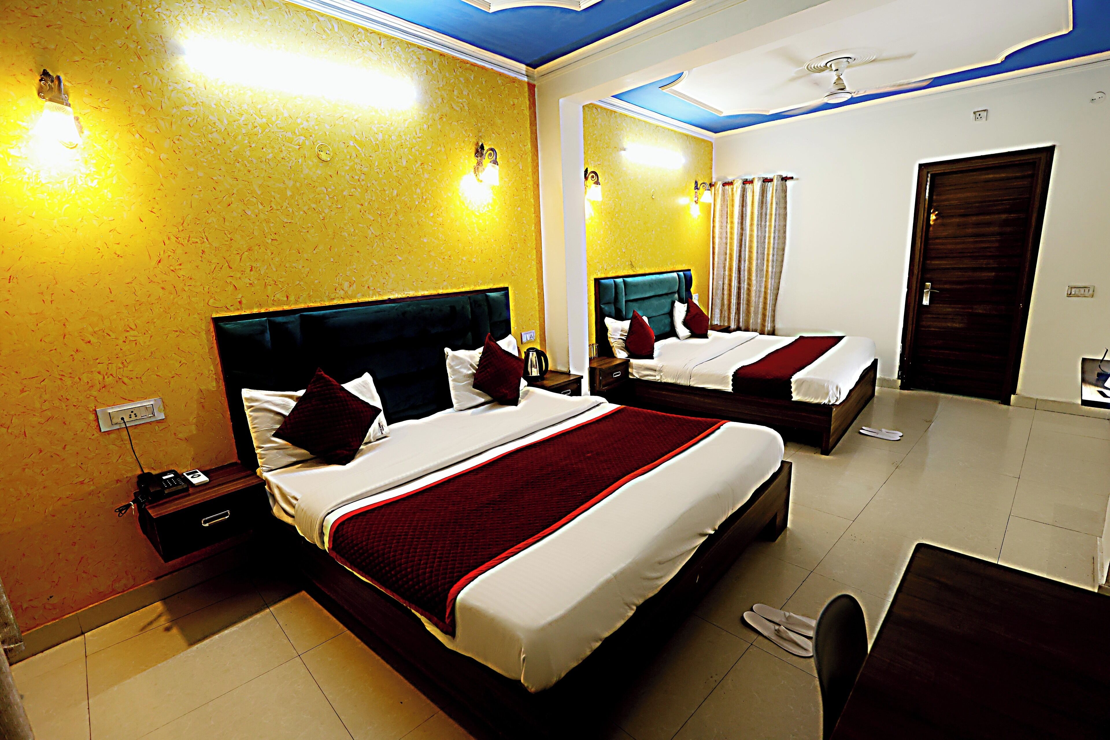 A hotel room with two beds, a desk, and a chair. There are bedside tables with lamps and a power outlet. The room has a ceiling fan and a door.