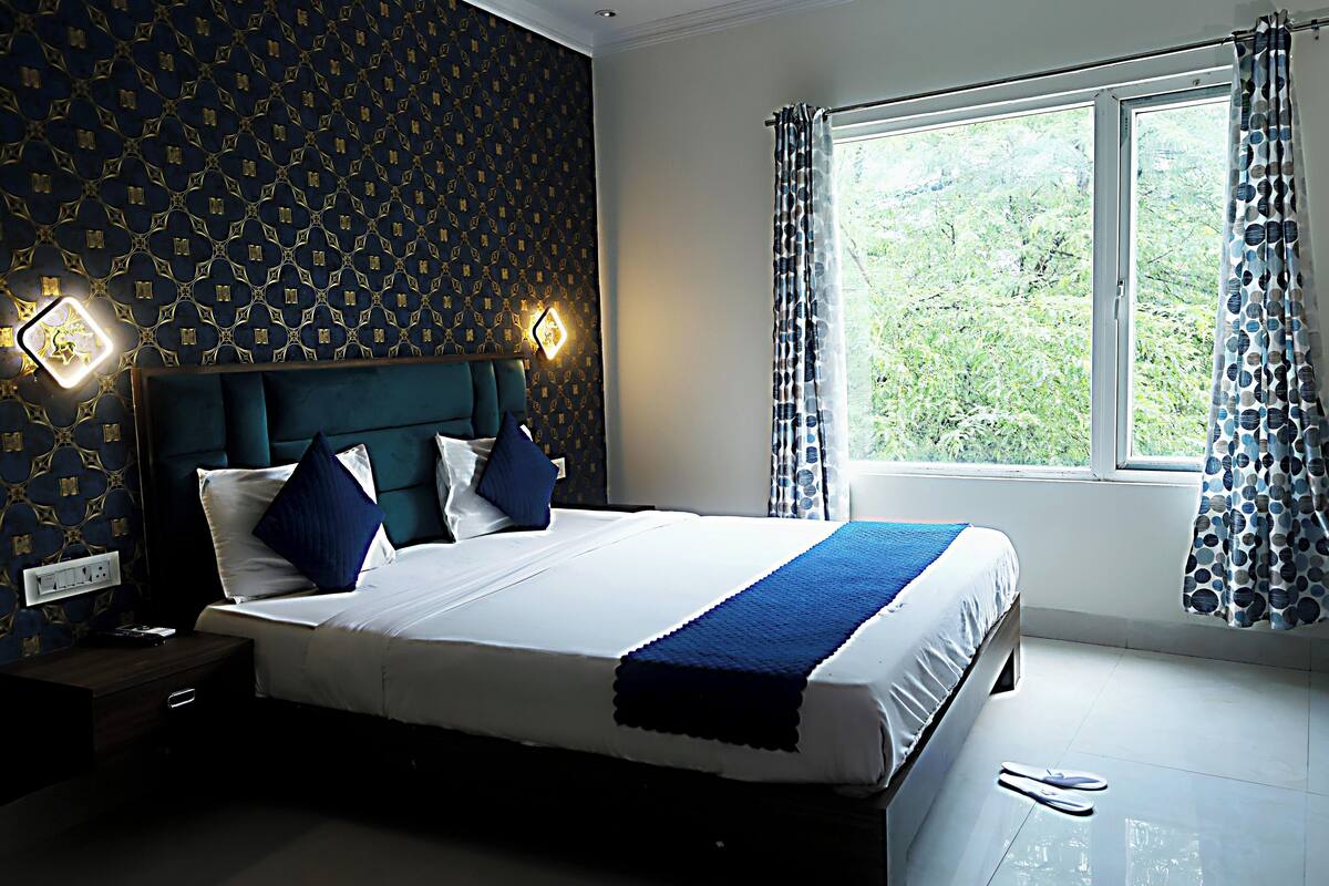 A neatly made bed with a blue accent, a nightstand with a lamp, and a window with curtains.