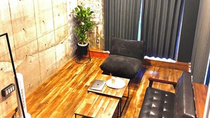 Un espacio habitable pequeño con suelo de madera, un sofá negro, una mesita, una planta en maceta y un aire acondicionado instalado en el techo.