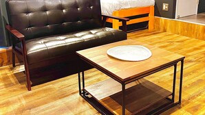 Una habitación con un sofá de cuero negro, una mesa de centro de madera con un posavasos blanco, una cama de madera con cabecera naranja y piso de madera.