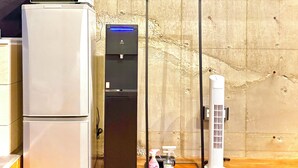 Una cocina con microondas, un refrigerador negro con dispensador de agua y un ventilador blanco.